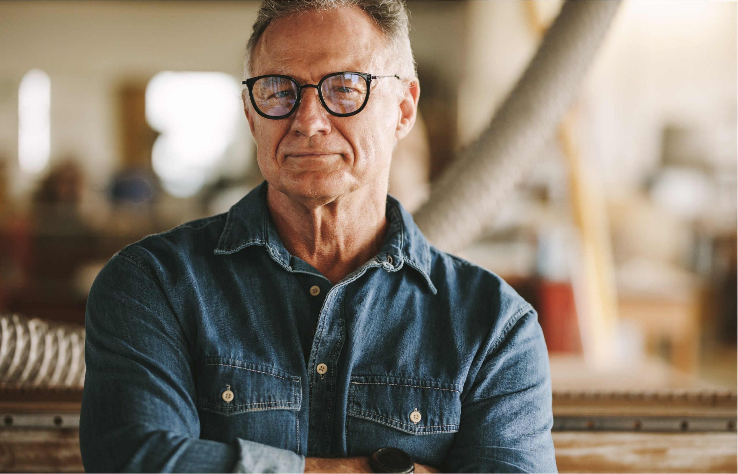 Portrait of successful senior man in eyeglasses standing in his carpentry workshop. Proud carpentry workshop owner standing with his arms crossed.