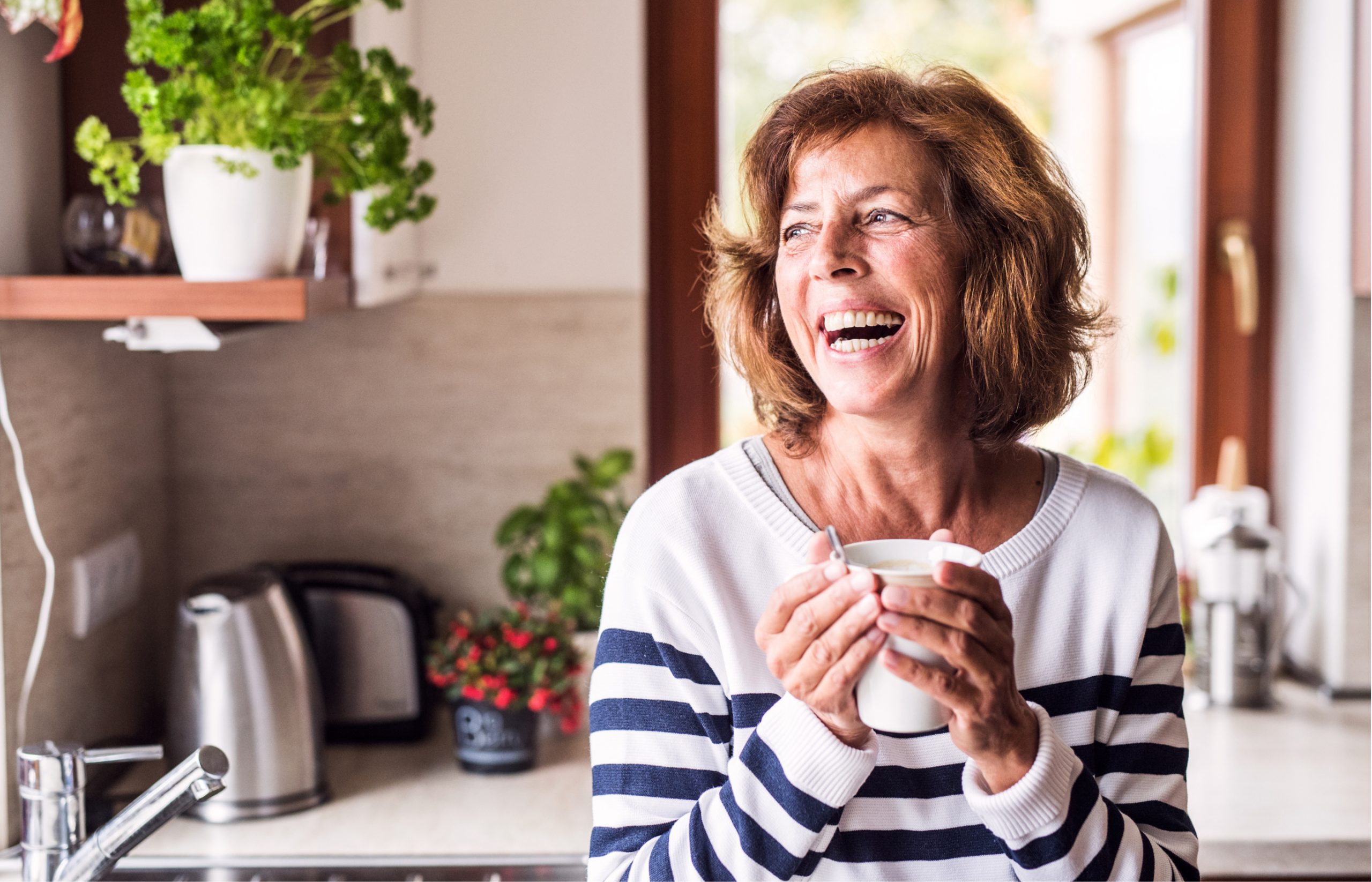 A high-net-worth retiree and wealth management client holds a coffee cup while laughing in her kitchen.