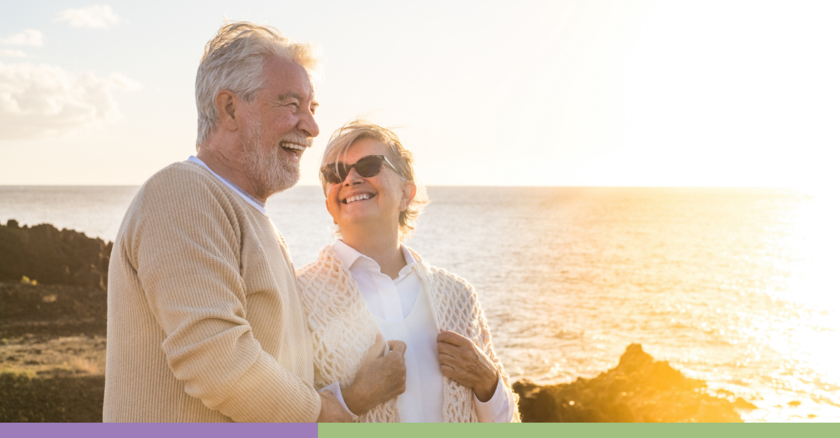A couple in retirement stares happily at each other alongside a sunny California coastline, with the ocean in the background.